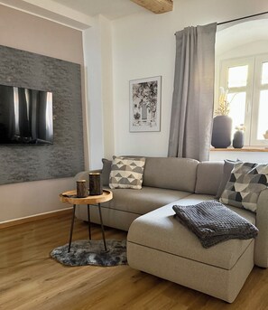 A modern living room with a grey sofa, a small wooden side table, and a grey rug. - Wohnzeit by Zirwick Apartment (Windischeschenbach)