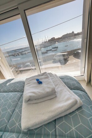 Ein Bett mit einem gefalteten Handtuch und einem blauen Gegenstand, mit Blick auf einen Jachthafen mit Booten und Gebäuden.
