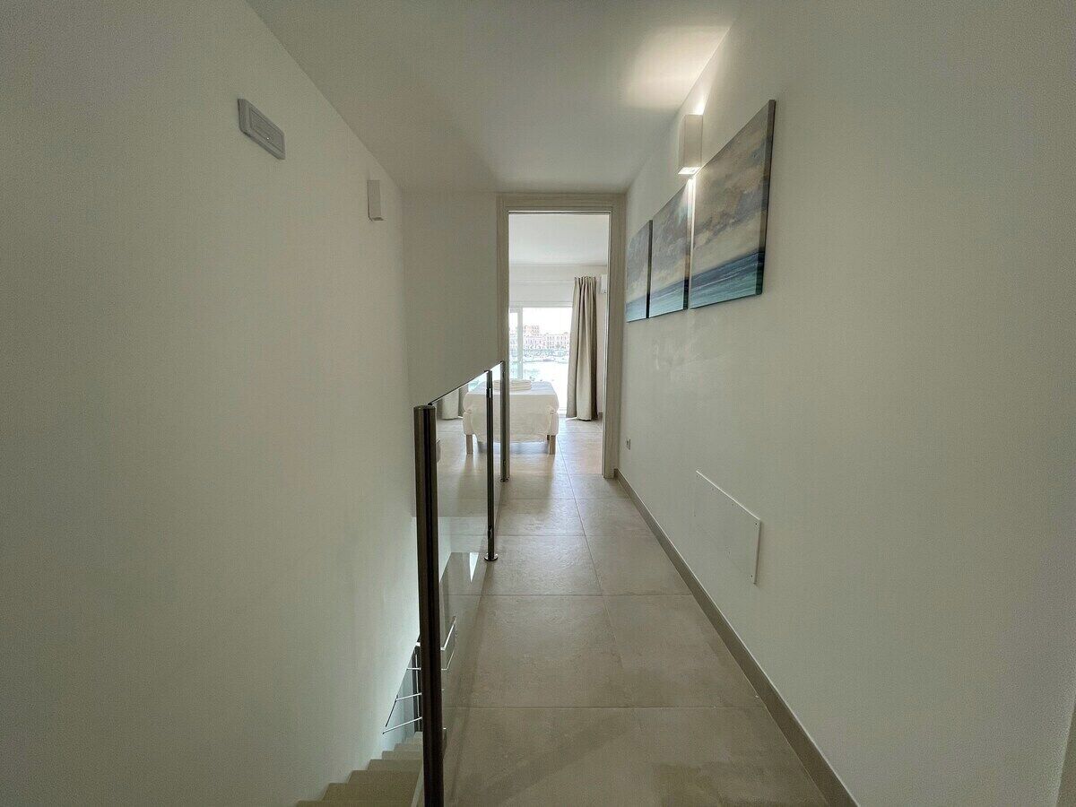 Un pasillo estrecho y bien iluminado con suelo de baldosas, una barandilla a la izquierda y un cuadro en la pared derecha.