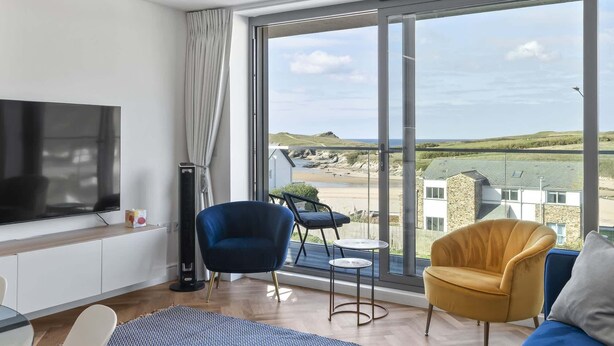 Ein modernes Wohnzimmer mit großem Fernseher, bequemen Sitzmöglichkeiten, einem kleinen Tisch und Blick auf Strand und Hügel.