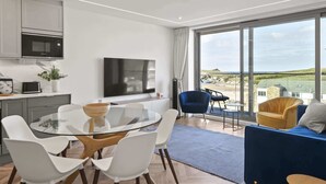 Ein modernes Wohnzimmer mit einem Glastisch, weißen Stühlen, einem blauen Sofa und einem großen Fenster mit Blick auf den Strand.