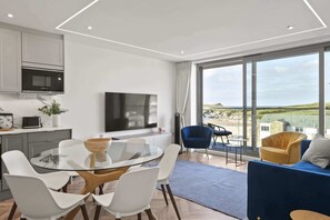 Ein modernes Wohnzimmer mit einem Glastisch, weißen Stühlen, einem blauen Sofa und einem großen Fenster mit Blick auf den Strand.