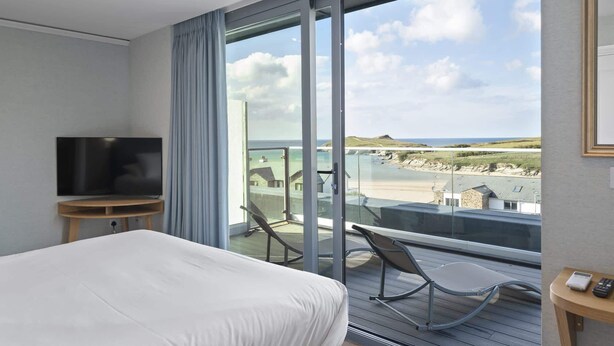 A hotel room with a bed, a TV, a balcony with a chair, and a view of the beach and ocean. - Penthouse, Balcony with Expansive Sea Views, Parking (Newquay)