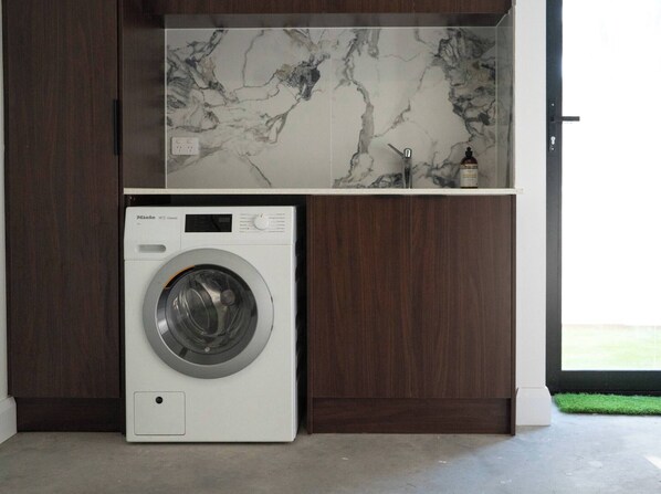 A modern laundry room with a white washing machine, dark wooden cabinets, and a marble backsplash. - Cosy Retreat Hilton SA (Adelaide)