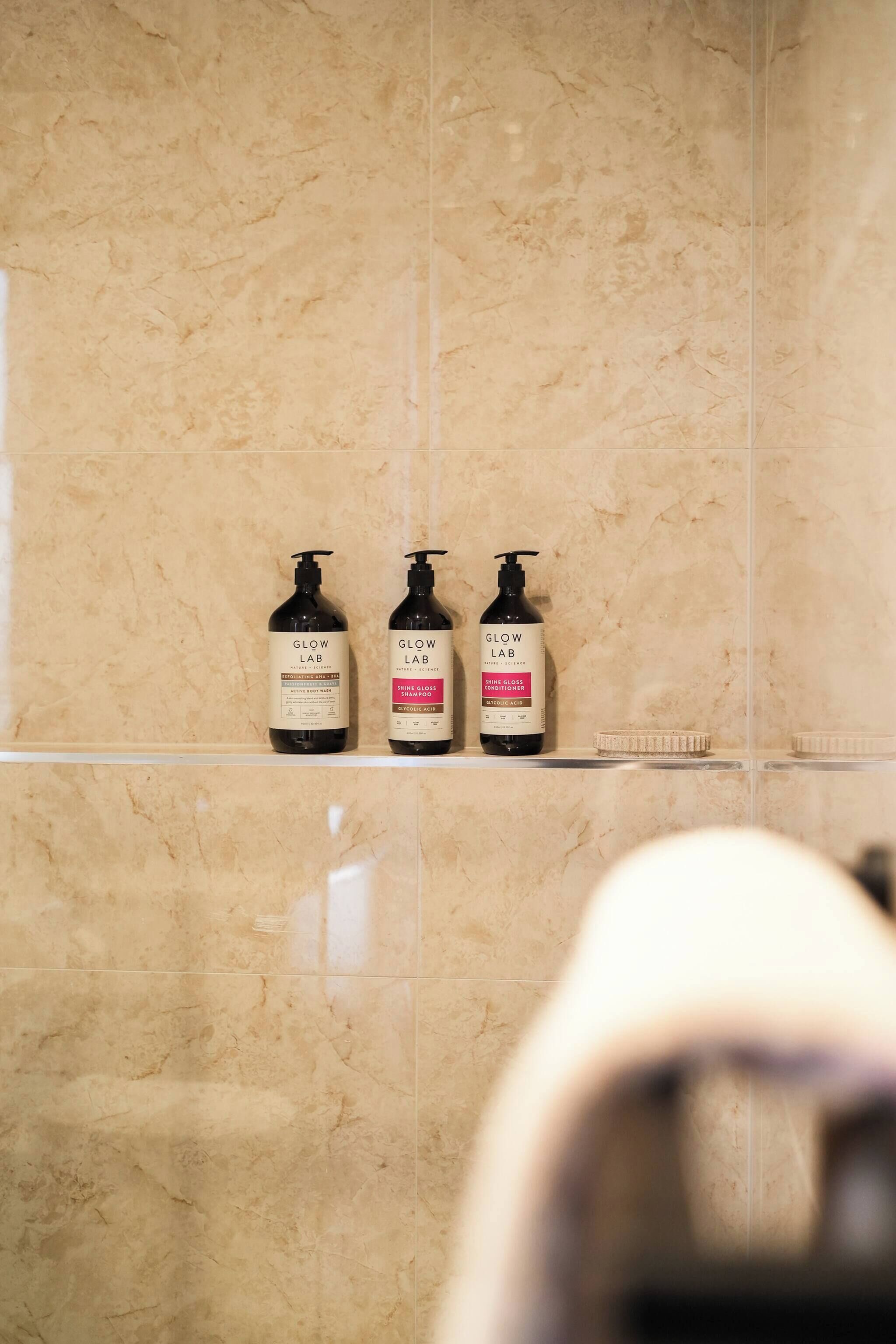 Ett badrum med tre flaskor schampo och balsam från Glow Lab på en hylla.