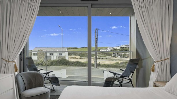 Una habitación de hotel moderna con un gran ventanal corredero que da a un balcón, dos sillas, una cama y vistas a la playa y a las casas.