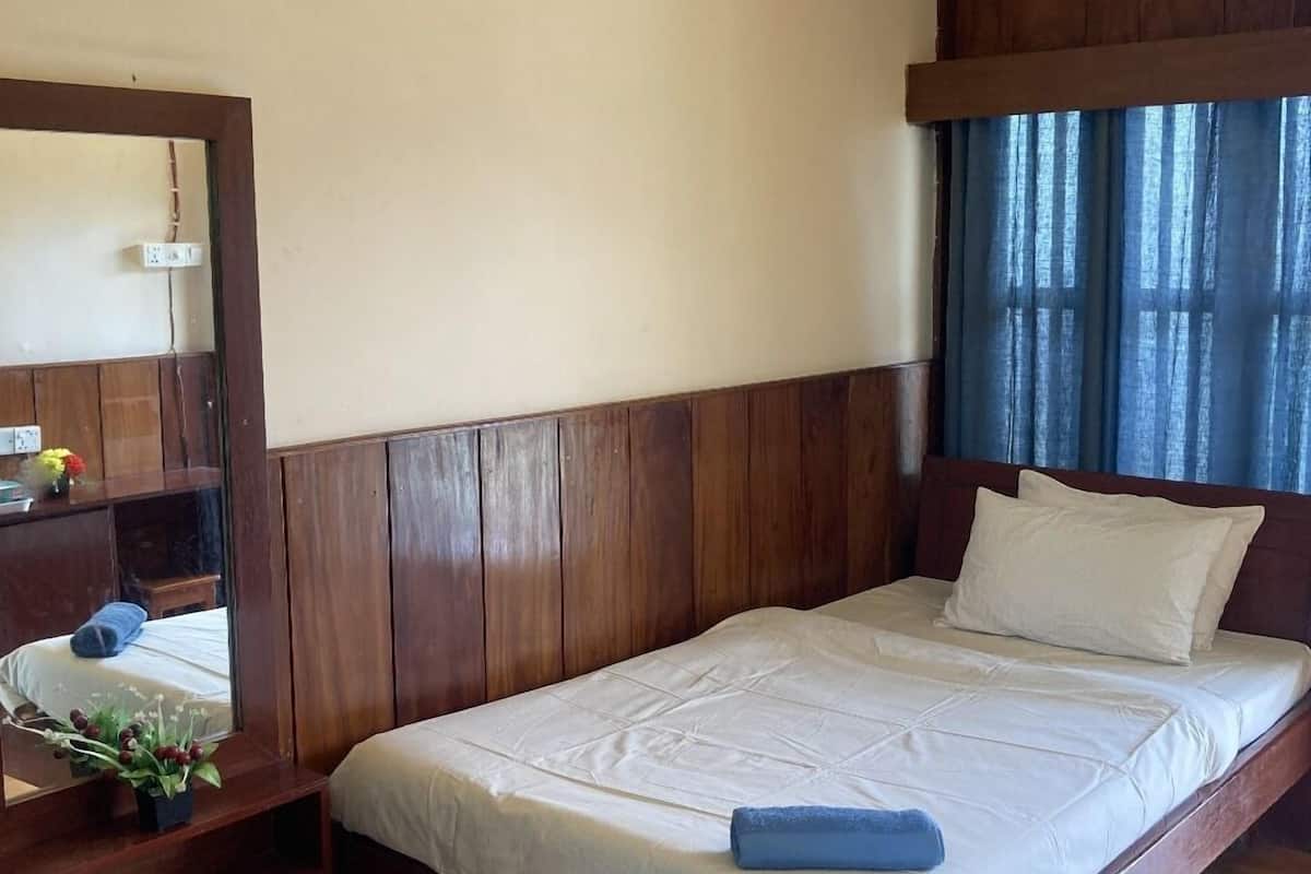 Una cama individual con estructura de madera y colchón blanco, ventilador de techo, espejo y ventana con cortinas.