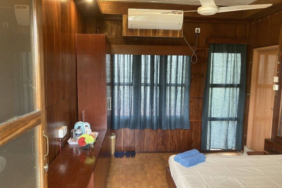 Una habitación con cama, escritorio con hervidor, ventana con cortinas y ventilador de techo.