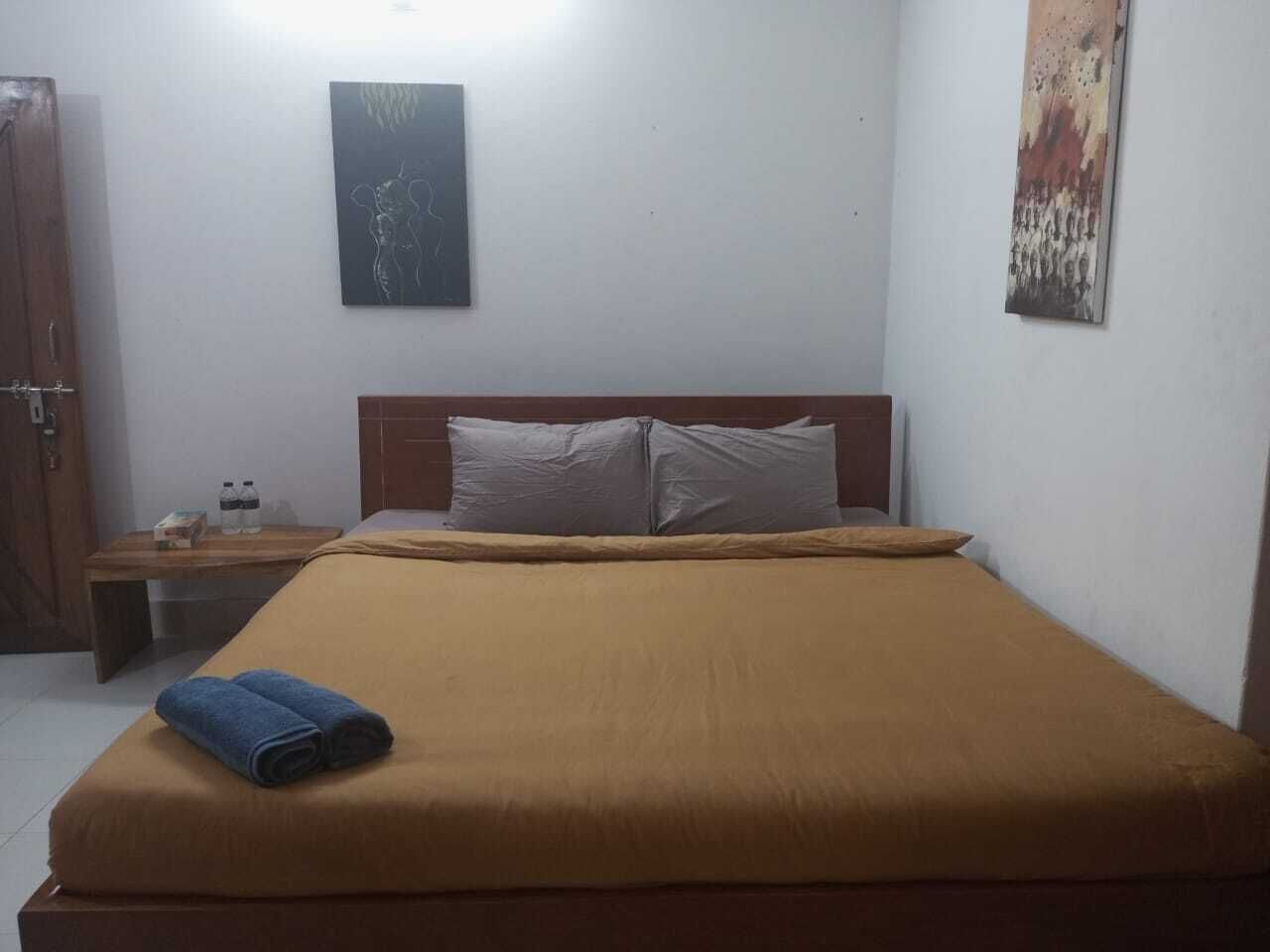 Una cama individual con una colcha marrón, un cabecero de madera y dos almohadas. Hay una mesita de noche de madera con una lámpara y una botella de agua. Se ve un cuadro en la pared sobre la cama.