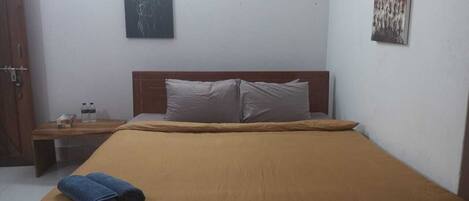 Una cama individual con una colcha marrón, un cabecero de madera y dos almohadas. Hay una mesita de noche de madera con una lámpara y una botella de agua. Se ve un cuadro en la pared sobre la cama.