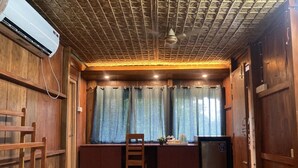 Ein kleines Zimmer mit Holzboden, einem Schreibtisch, einem Stuhl, einem Vorhangfenster und einer Wandklimaanlage.