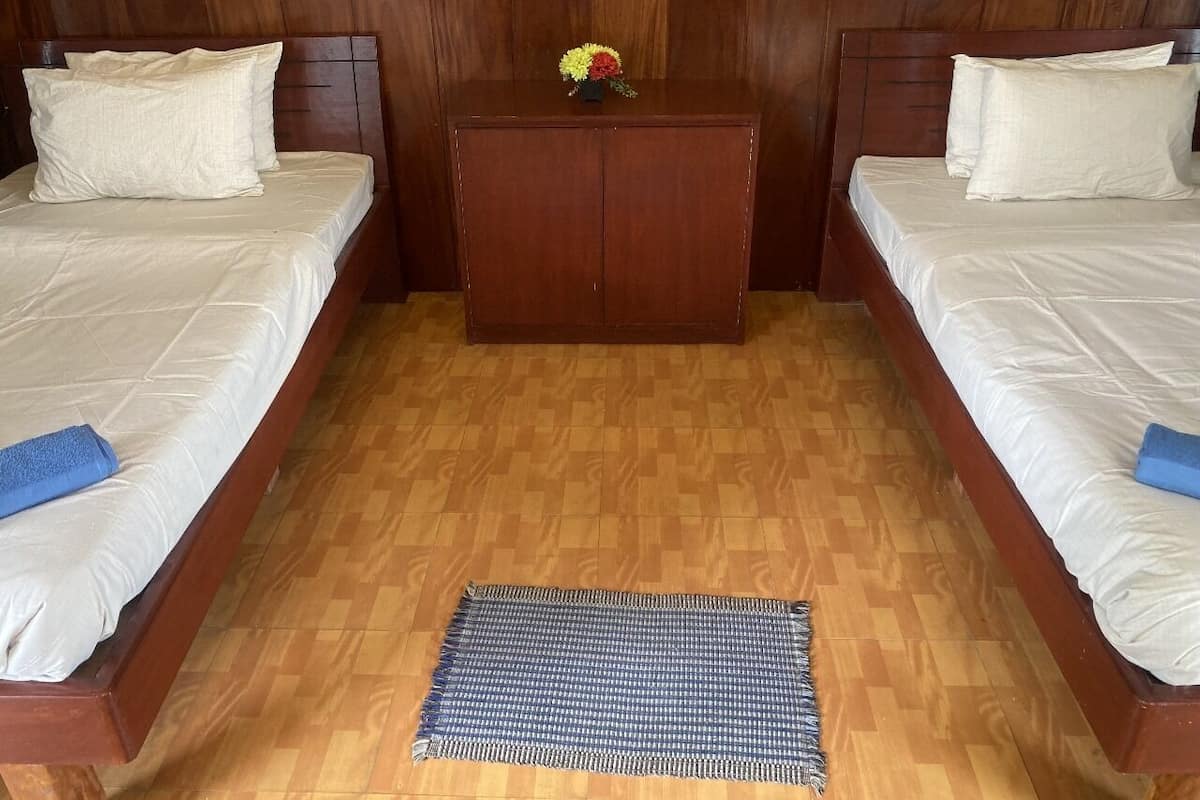 Una habitación con dos camas, cabeceros de madera, una mesita pequeña con un florero y un tapete a cuadros en el suelo.