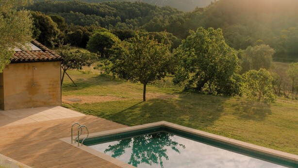 A swimming pool with a wooden deck, surrounded by a grassy area and trees, with a mountain in the background. - Can Terrades by Artu (Beuda)