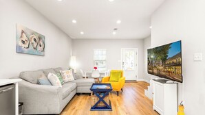 A modern living room with a grey sofa, a yellow armchair, a blue coffee table, and a flat-screen TV on a white stand. - 3 Bedrooms | Downtown Pensacola | Walkable Location | Fenced Yard (Pensacola)