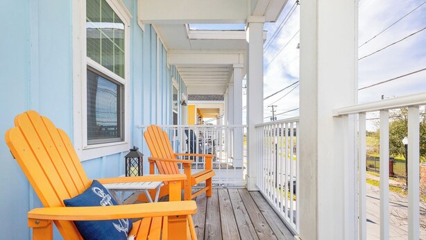 A porch with wooden flooring and white railings, featuring two orange Adirondack chairs and a small table with a lantern. - 3 Bedrooms | Downtown Pensacola | Walkable Location | Fenced Yard (Pensacola)