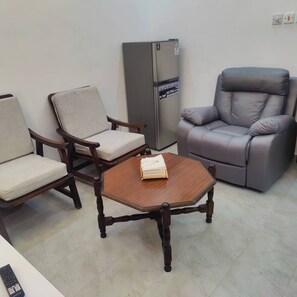 Ein Zimmer mit einem Holztisch, zwei Stühlen, einem grauen Ledersessel und einem Kühlschrank.
