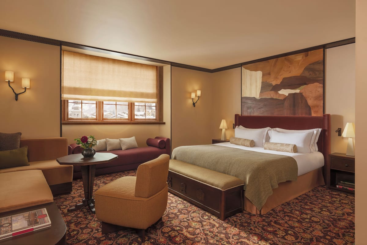 A hotel room with a large bed, a sofa, a chair, a small table with a vase, and a window with a view of a building.