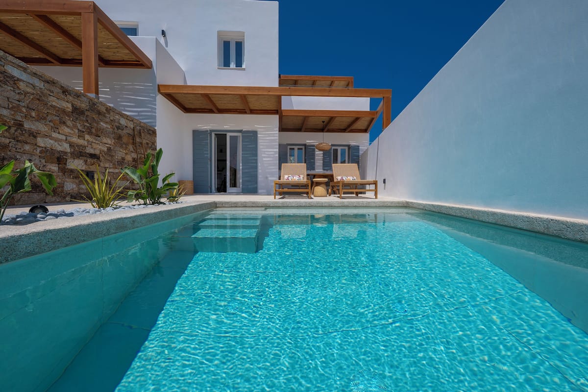 A modern pool area with a clear blue pool, wooden deck, and a white building with a stone wall.