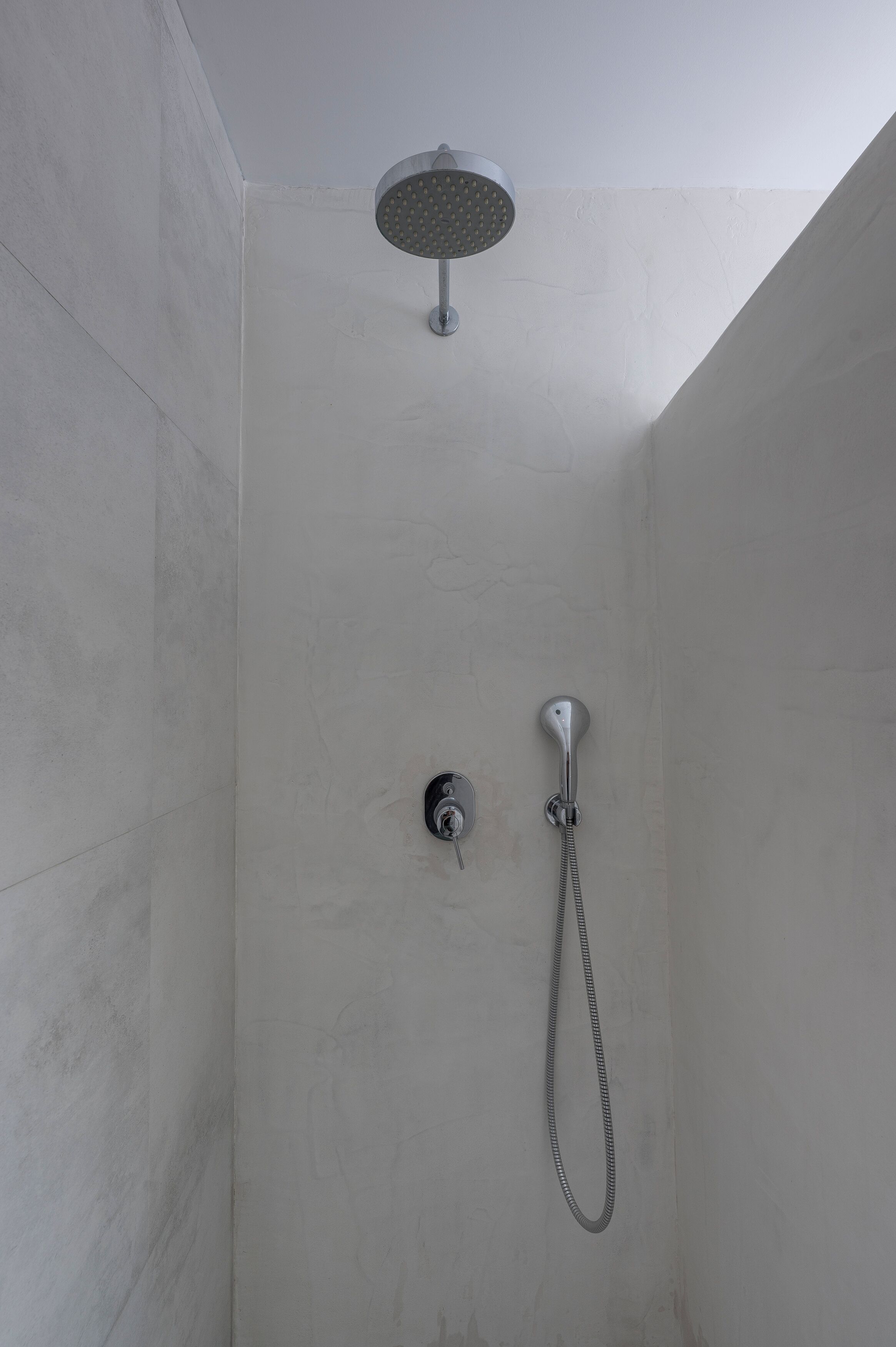 Una ducha con cabezal de mano y grifo empotrado en la pared, ubicada en un rincón de una habitación de paredes lisas.