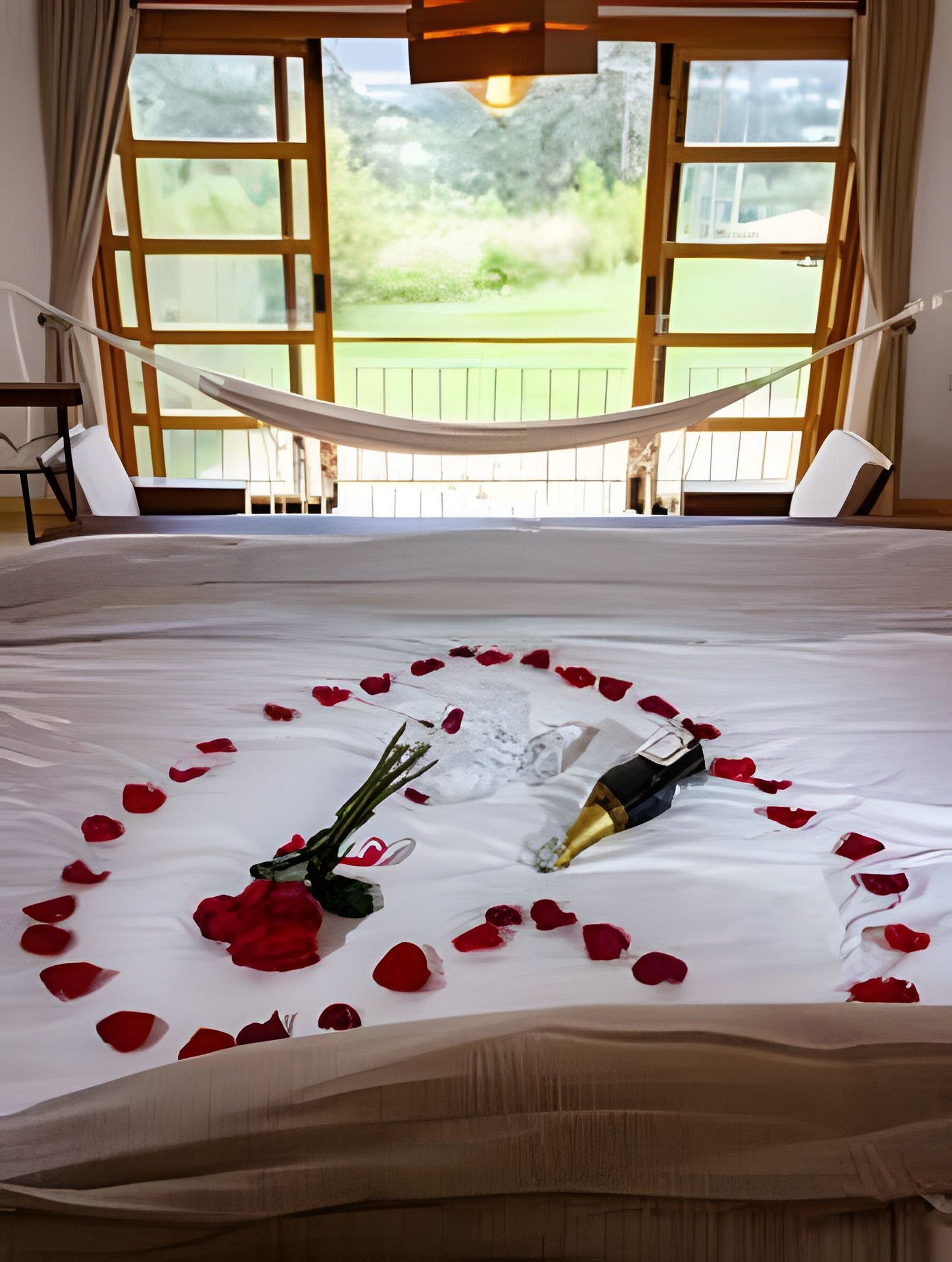 Ein Bett mit Rosenblättern und einer Champagnerflasche, eine Hängematte und Blick ins Grüne durch große Fenster.