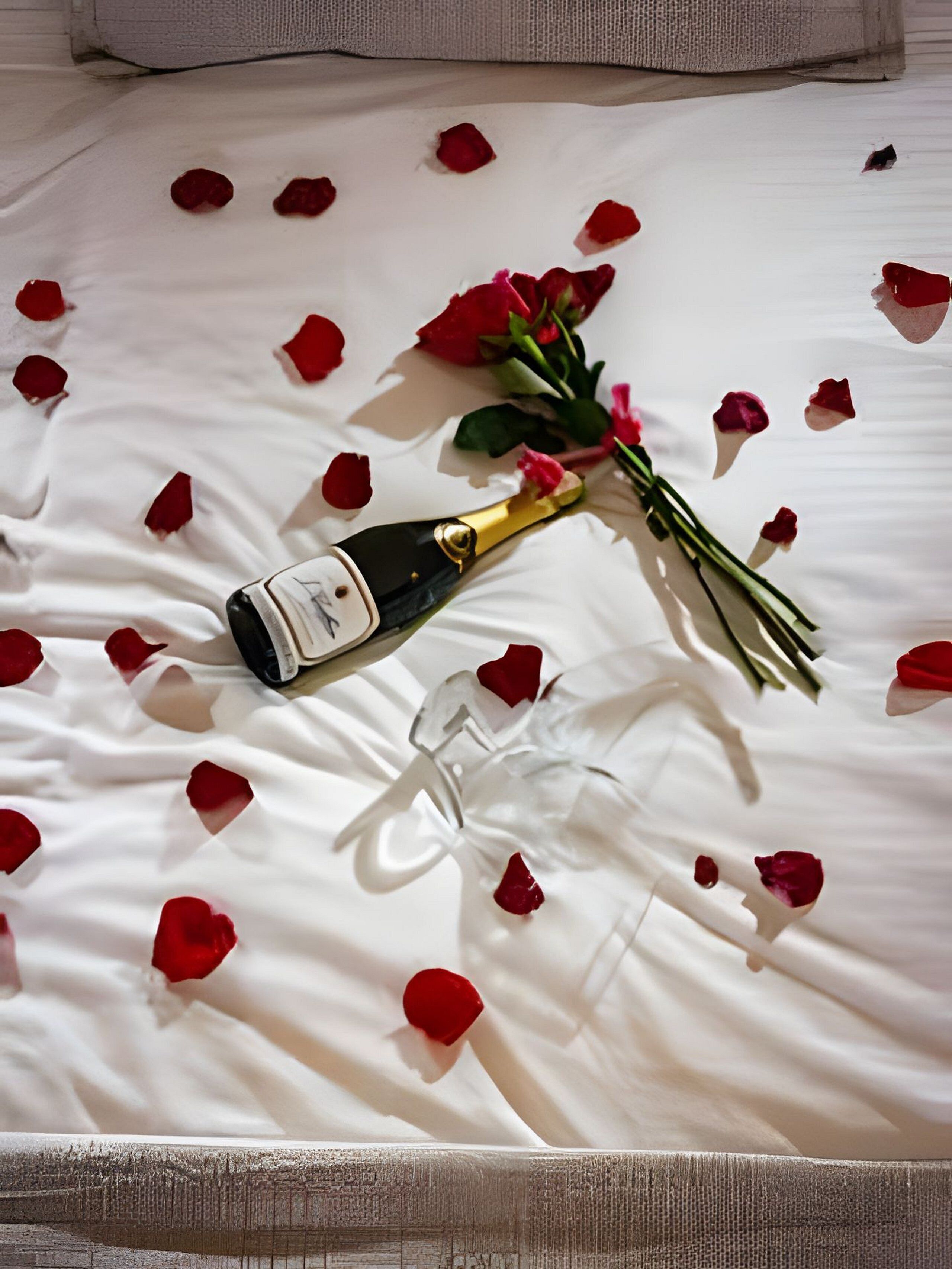 A bed with white sheets, a bouquet of red roses, and a bottle of champagne.