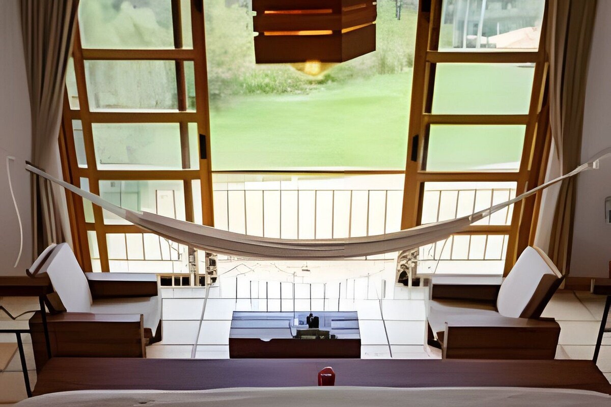 Un dormitorio con una cama grande, una hamaca y vistas a un campo de golf a través de ventanas abiertas.