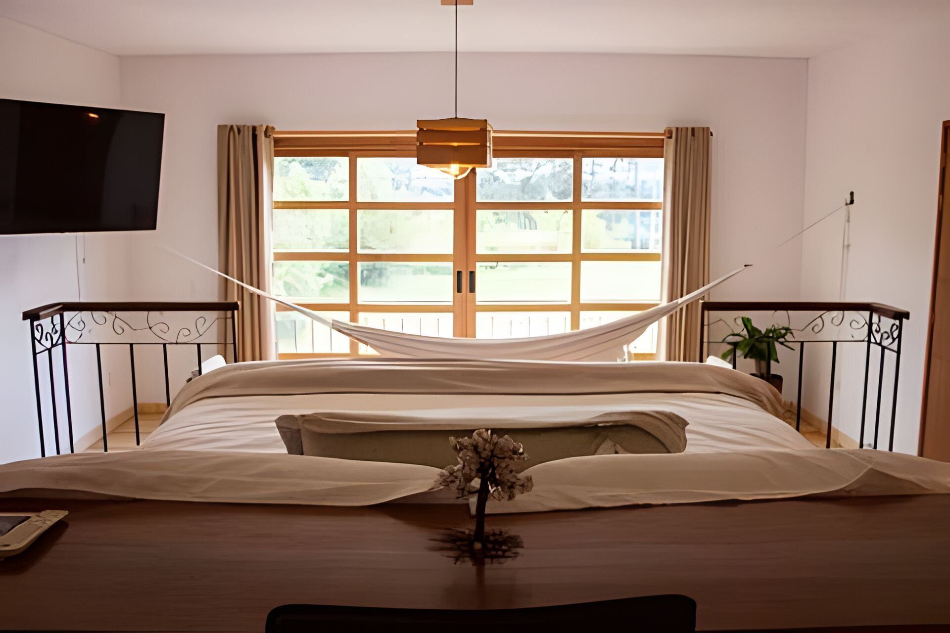 A neatly made bed with a hammock, a potted plant, and a TV mounted on the wall.