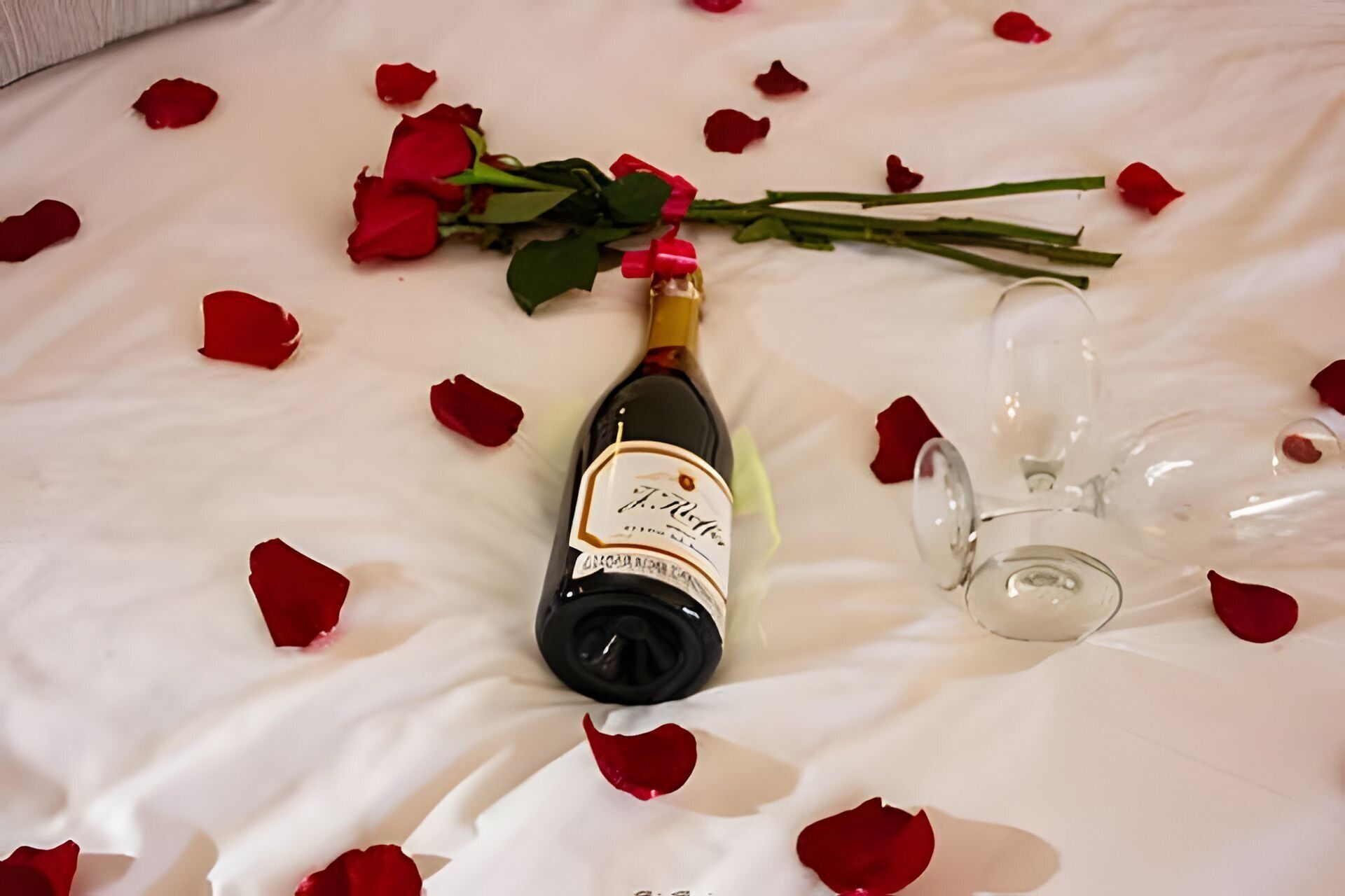 Uma garrafa de vinho, um copo e pétalas de rosa sobre uma superfície branca.