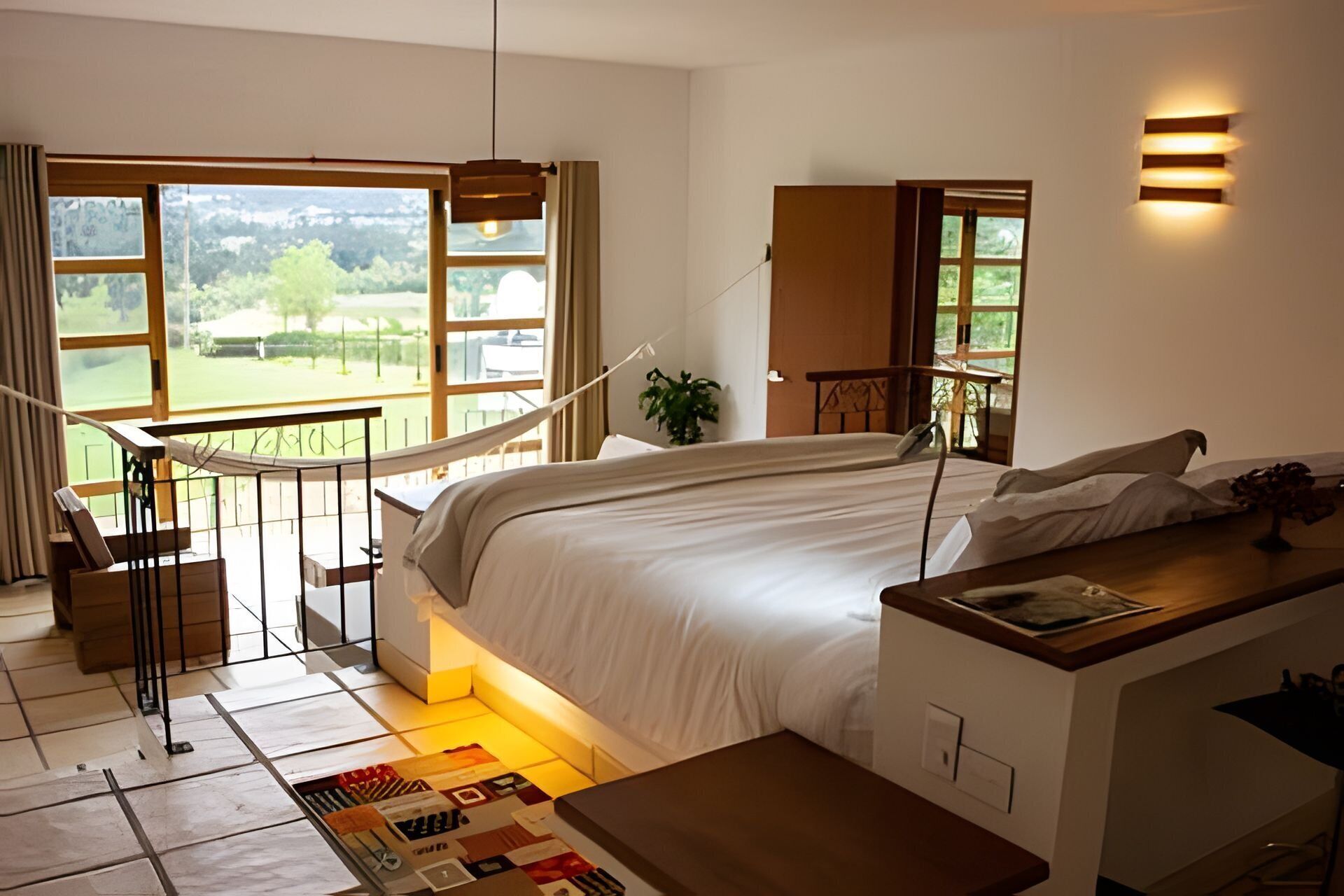 A bedroom with a large bed, a desk, a hammock, and a view of a green landscape.