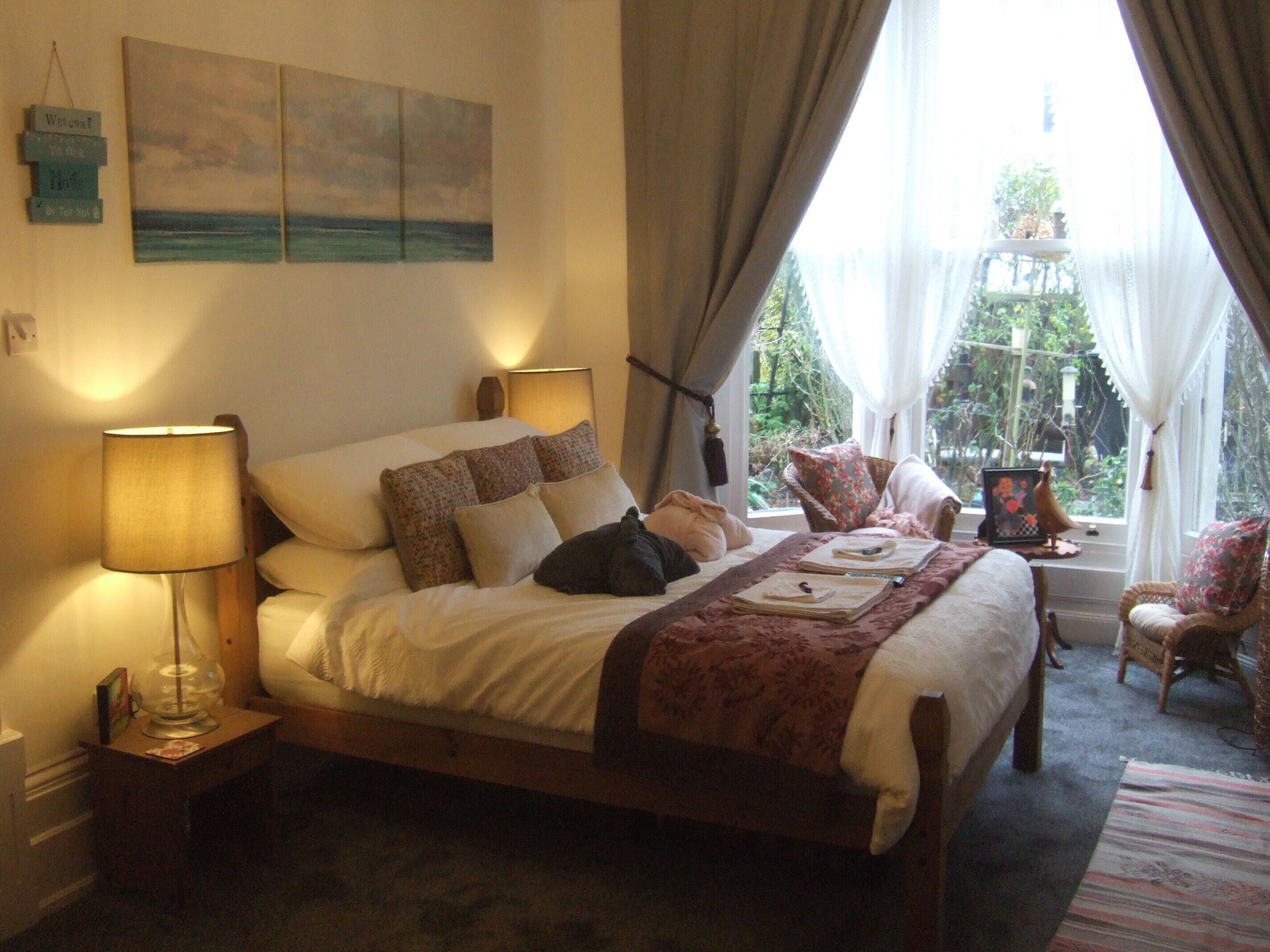 A bedroom with a bed, bedside lamps, a window with curtains, and a view of greenery outside.