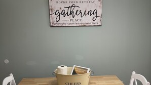 A dining area with a wooden table, white chairs, a sign on the wall, and a bucket with a card.