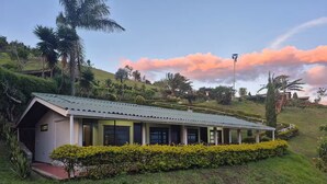 Un bâtiment d’un seul étage, doté d’un toit végétalisé et d’une allée couverte, entouré d’une végétation luxuriante et de collines.