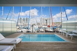 A pool area with a view of a marina, white lounge chairs, and a wooden deck.