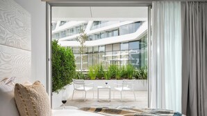 A modern bedroom with a large bed, a view of a glass building, and a balcony with chairs and plants. - The YACHT – Limited Edition By Fattal (Herzliya)