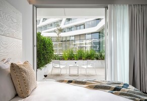 A modern bedroom with a large bed, a view of a glass building, and a balcony with chairs and plants.