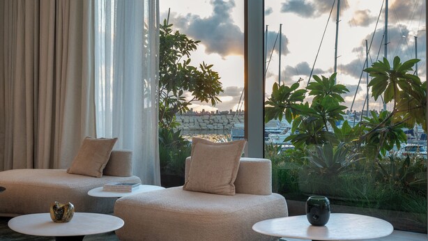 A modern living area with a sofa, two round tables, and a view of boats and greenery. - The YACHT – Limited Edition By Fattal (Herzliya)