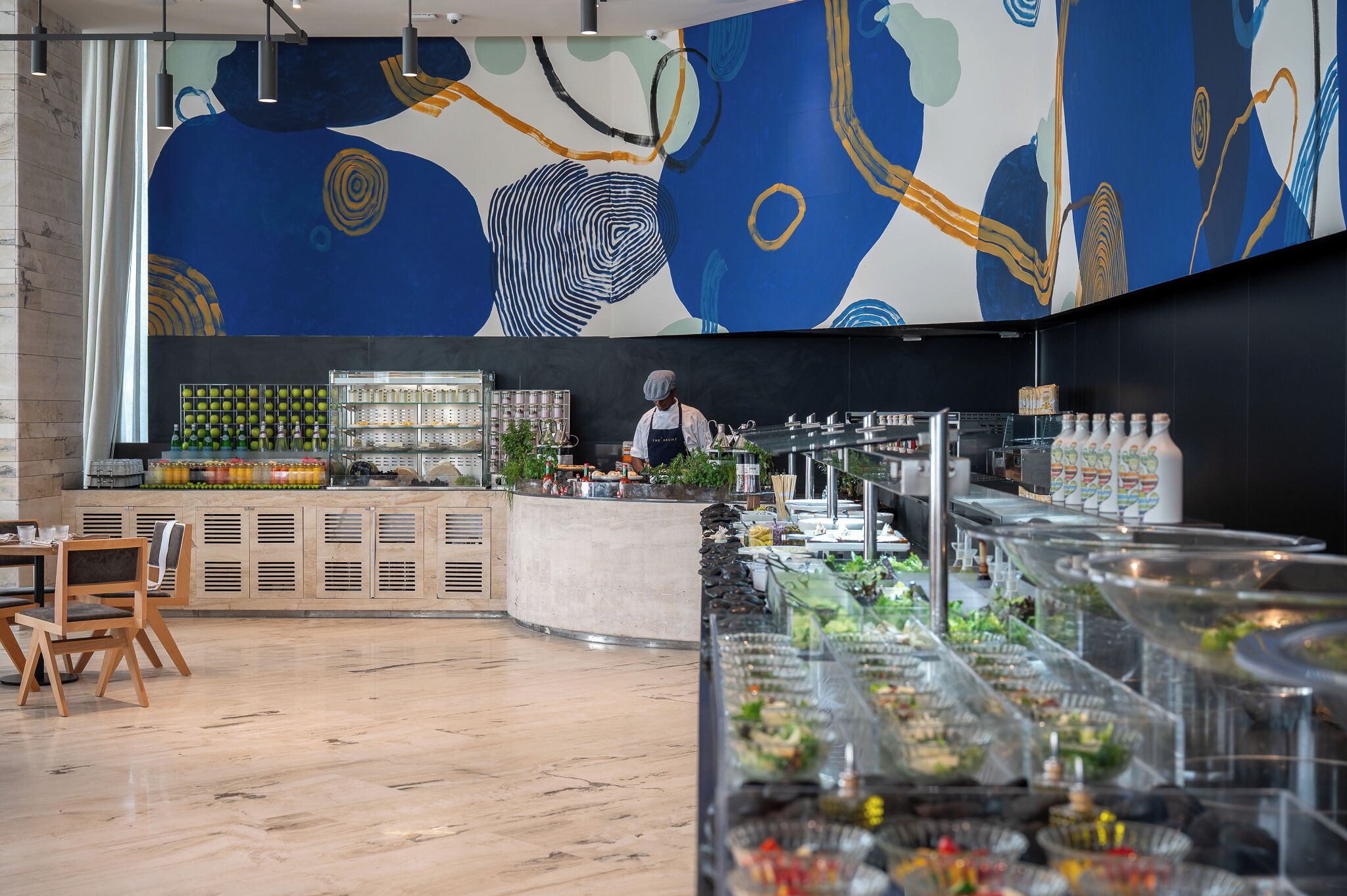Uma área de buffet com um chef preparando comida, um balcão com bebidas e uma parede com arte abstrata.