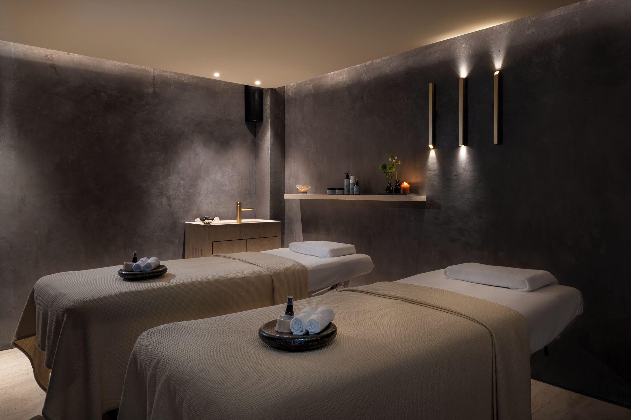 A spa room with two massage tables, a small vanity, and wall-mounted lighting.