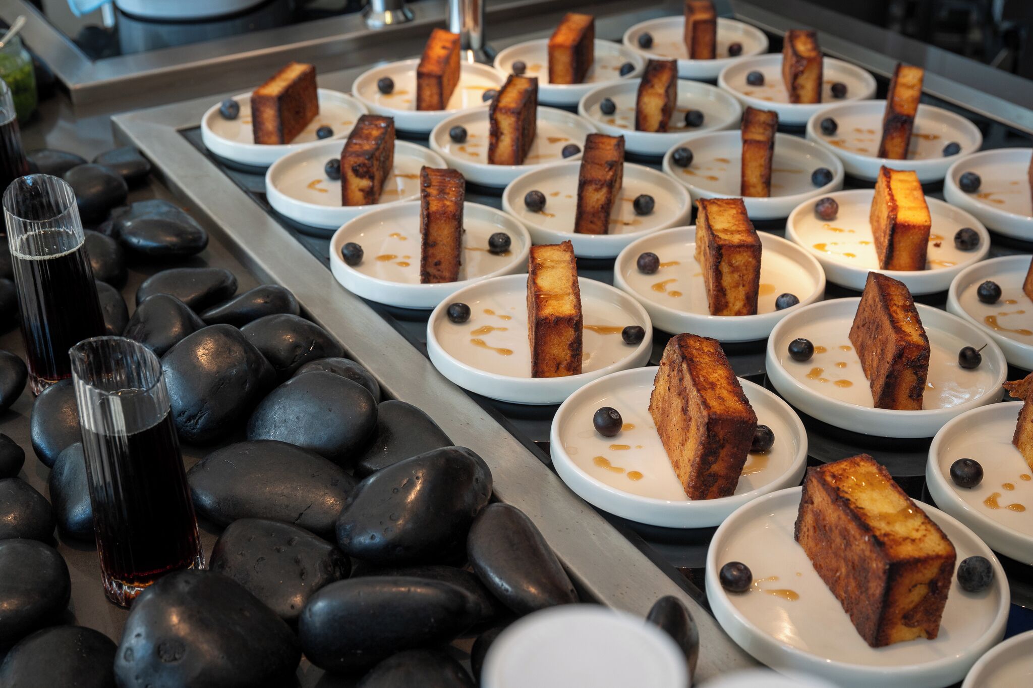 Een buffet met rechthoekige etenswaren op borden, gerangschikt op een dienblad met zwarte kiezelstenen en heldere glazen met een donker drankje.