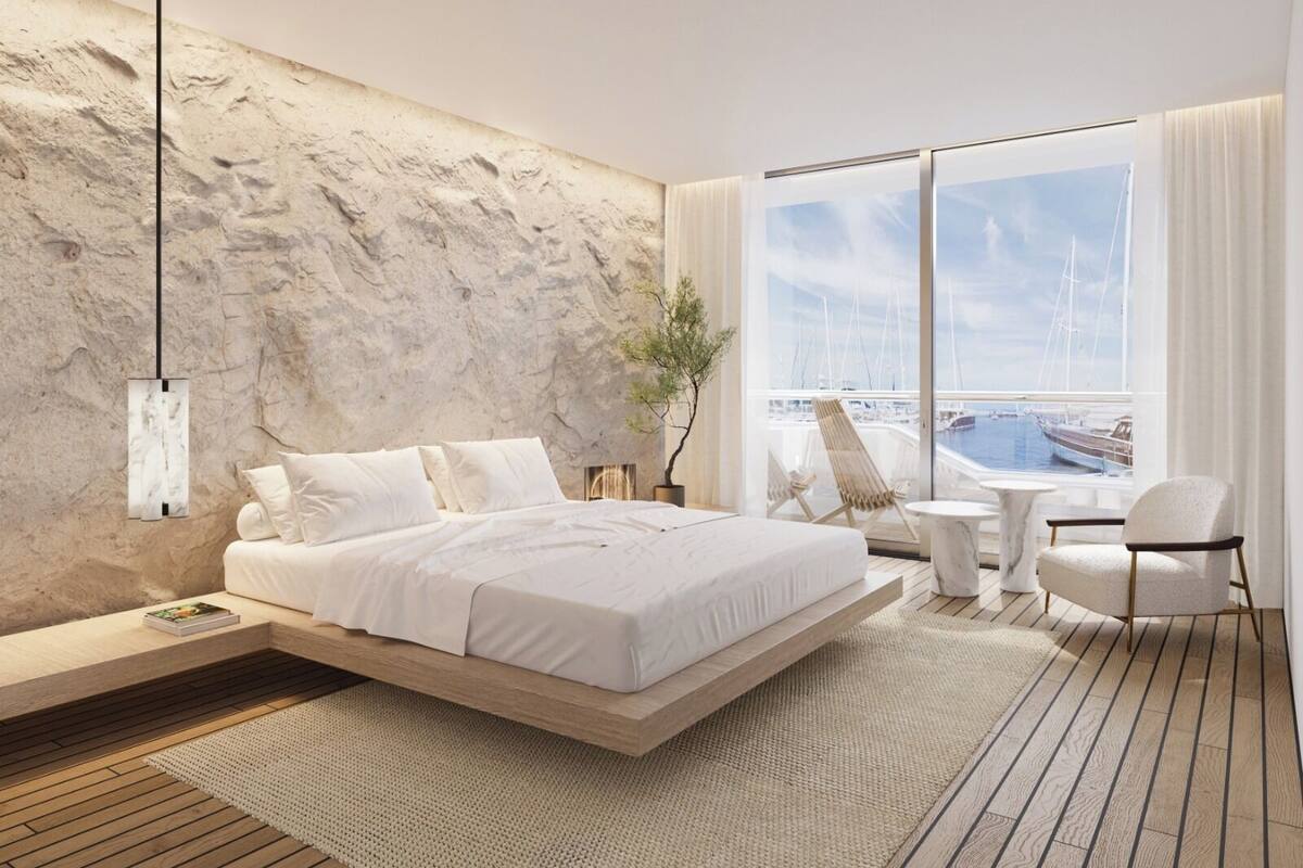 A modern bedroom with a bed, a side table, a chair, and a view of a marina through large windows.