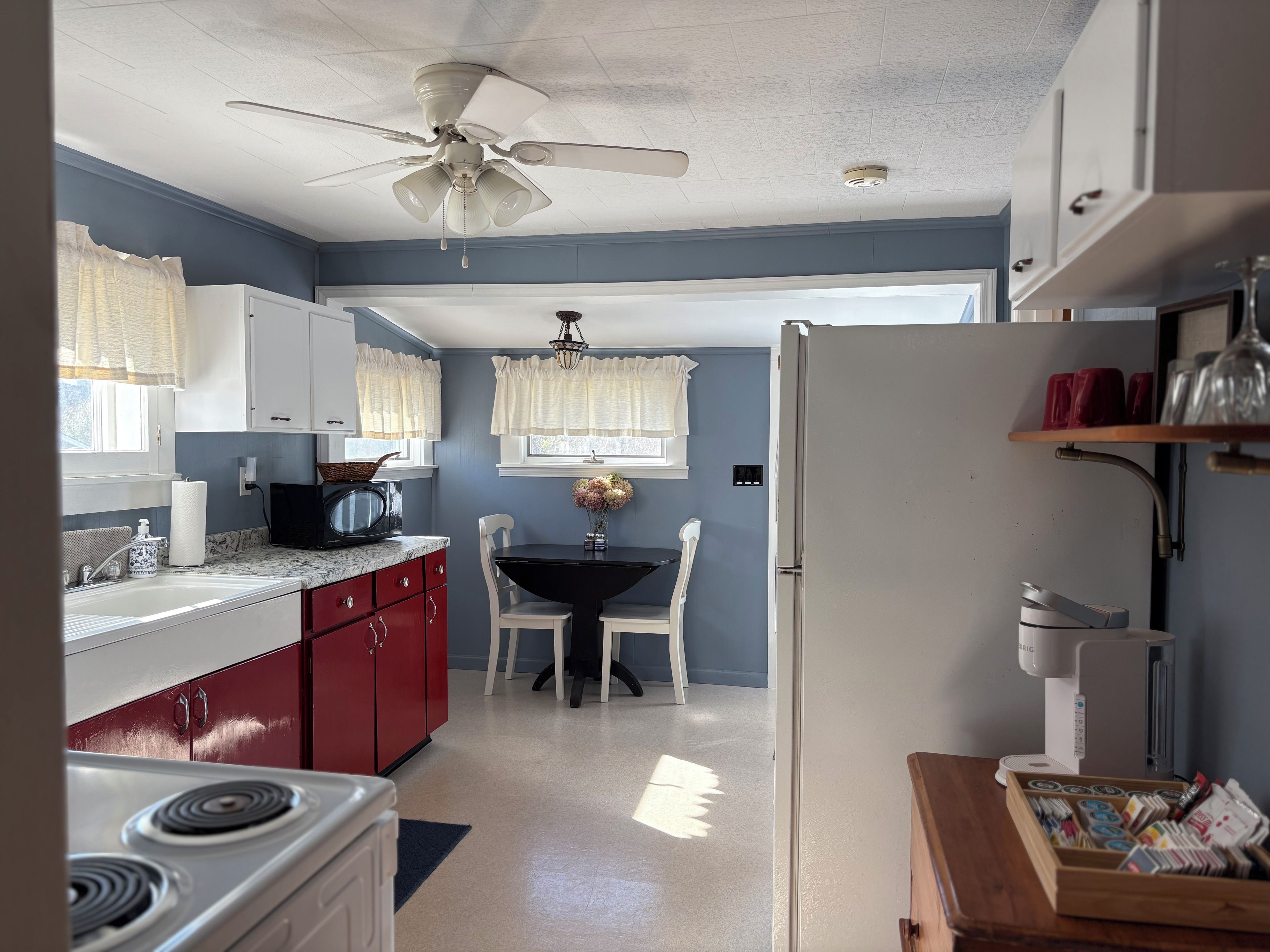 Une cuisine équipée d’une cuisinière, d’un four à micro-ondes et de meubles de rangement rouges. On y trouve une table à manger avec des chaises et un ventilateur de plafond.
