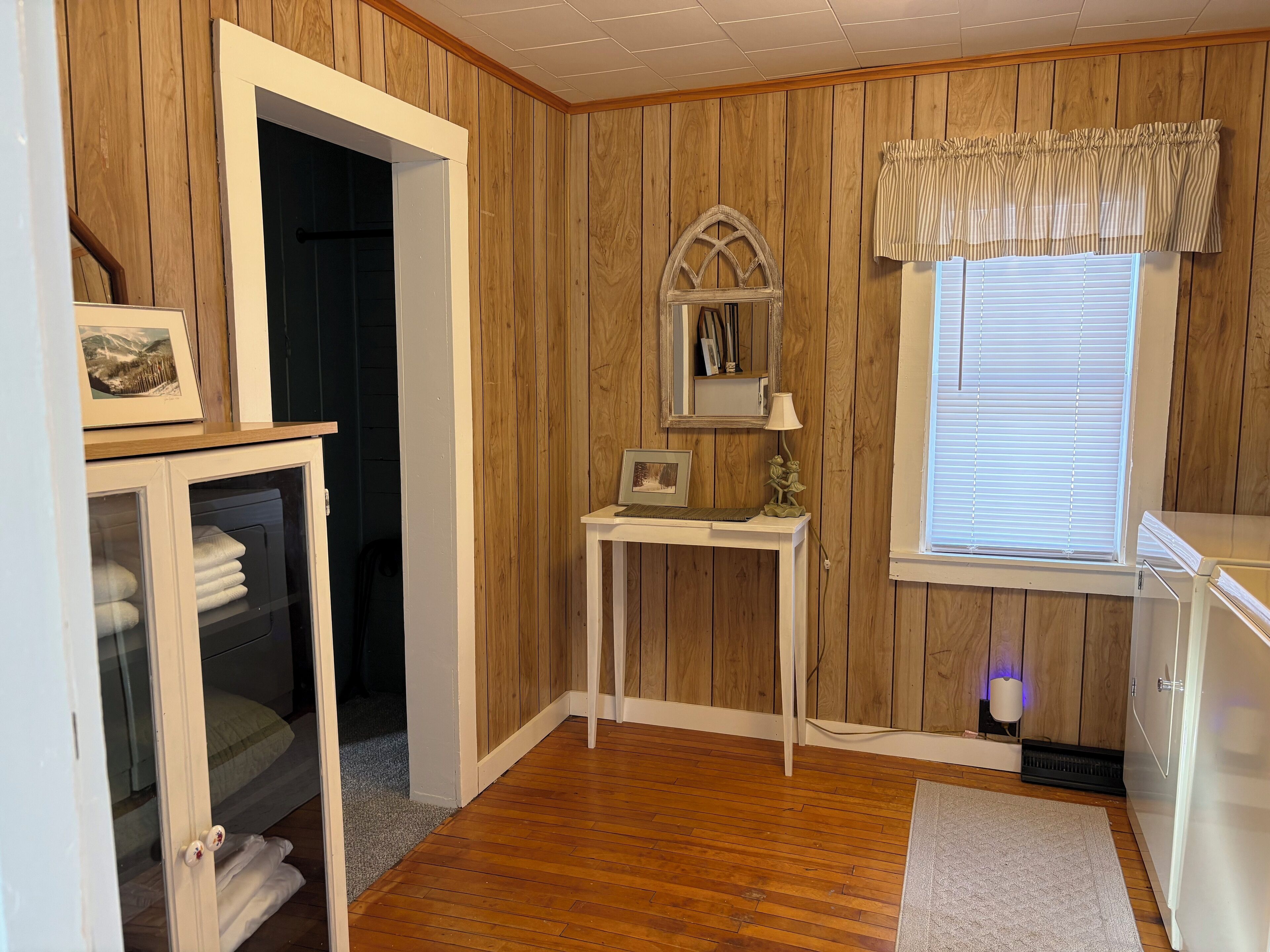 A room with wooden panel walls, a white door, a small table with a lamp, a window with blinds, and a white cabinet with towels.
