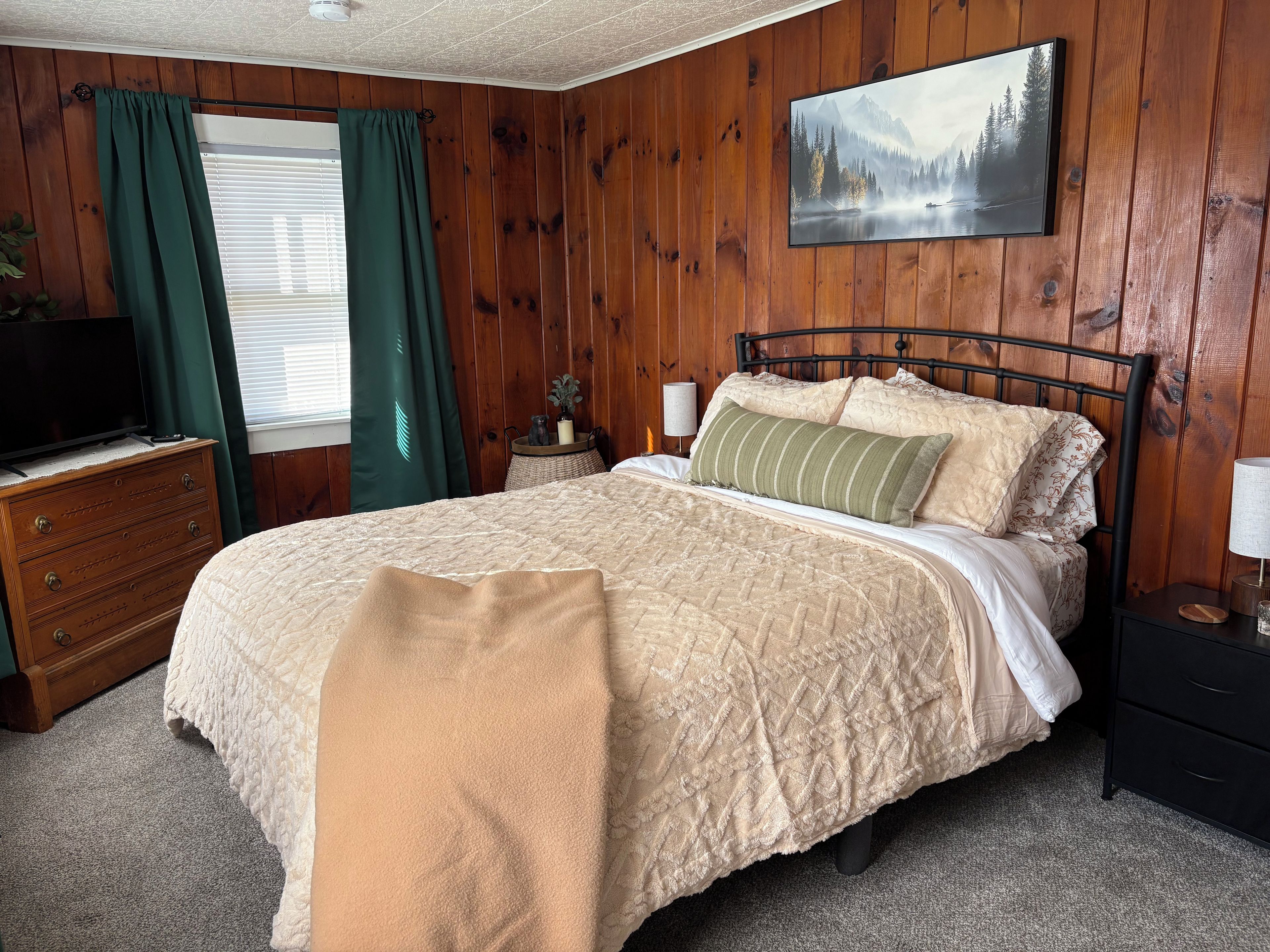 Un dormitorio con cama, cómoda, televisor, cuadro y ventana con cortinas.