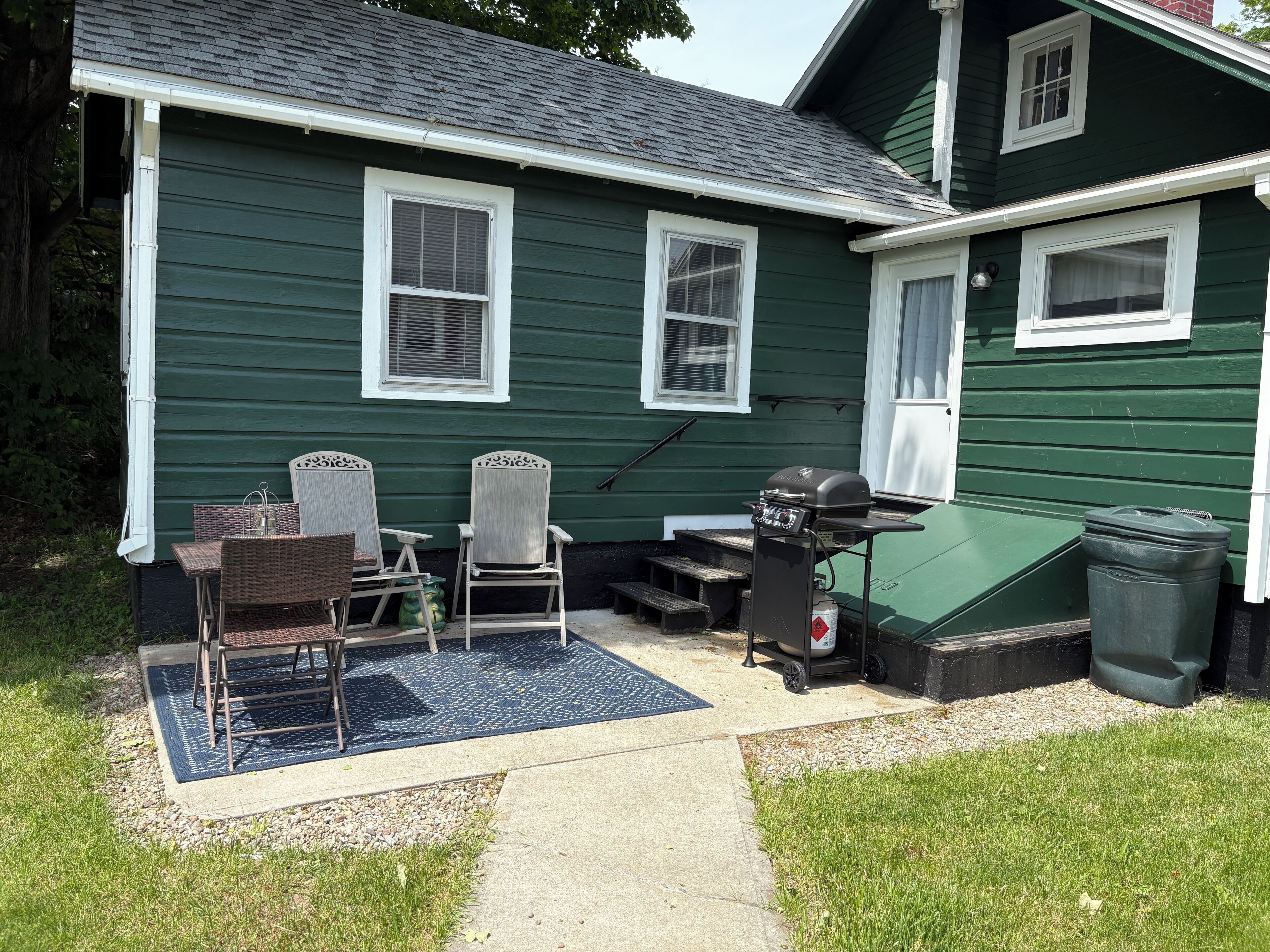Ein grünes Haus mit Terrasse, Sitzgelegenheiten im Freien, Grill und Mülleimer.