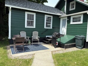 A green house with a patio, outdoor seating, a grill, and a trash can.
