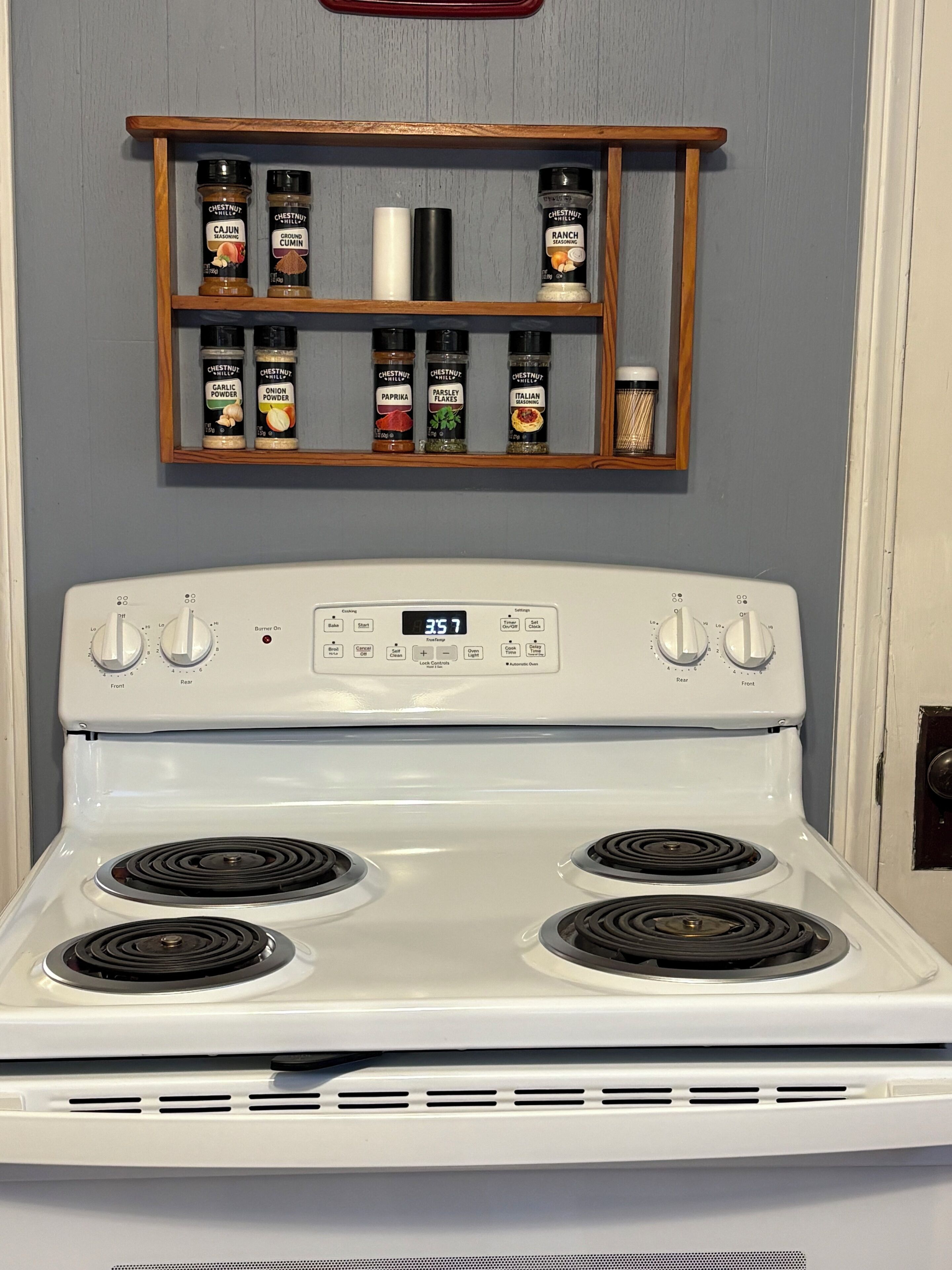 A white stove with four burners and a digital clock displaying 3:57. Above the stove, there is a wooden spice rack with various seasonings.
