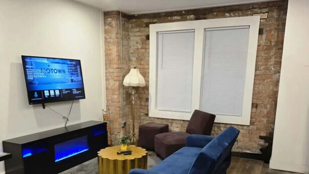 A modern living room with a blue sofa, two gold round coffee tables, a TV mounted on the wall, and a brick accent wall. - Spacious 6-bedroom apartment with AC in peaceful Detroit (Detroit)