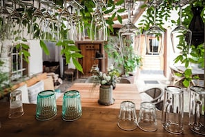 A wooden table with various glassware, including wine glasses and tumblers, in a cozy, plant-filled indoor setting. - Kaffee&Kissen - Boutique Hotel a. Rhein (Oestrich-Winkel)