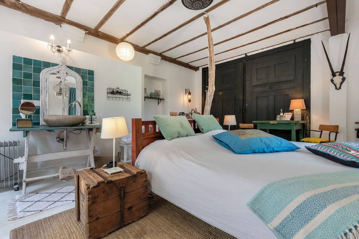 A bedroom with a bed, bedside tables, a mirror, a chandelier, and a rustic wooden chest.