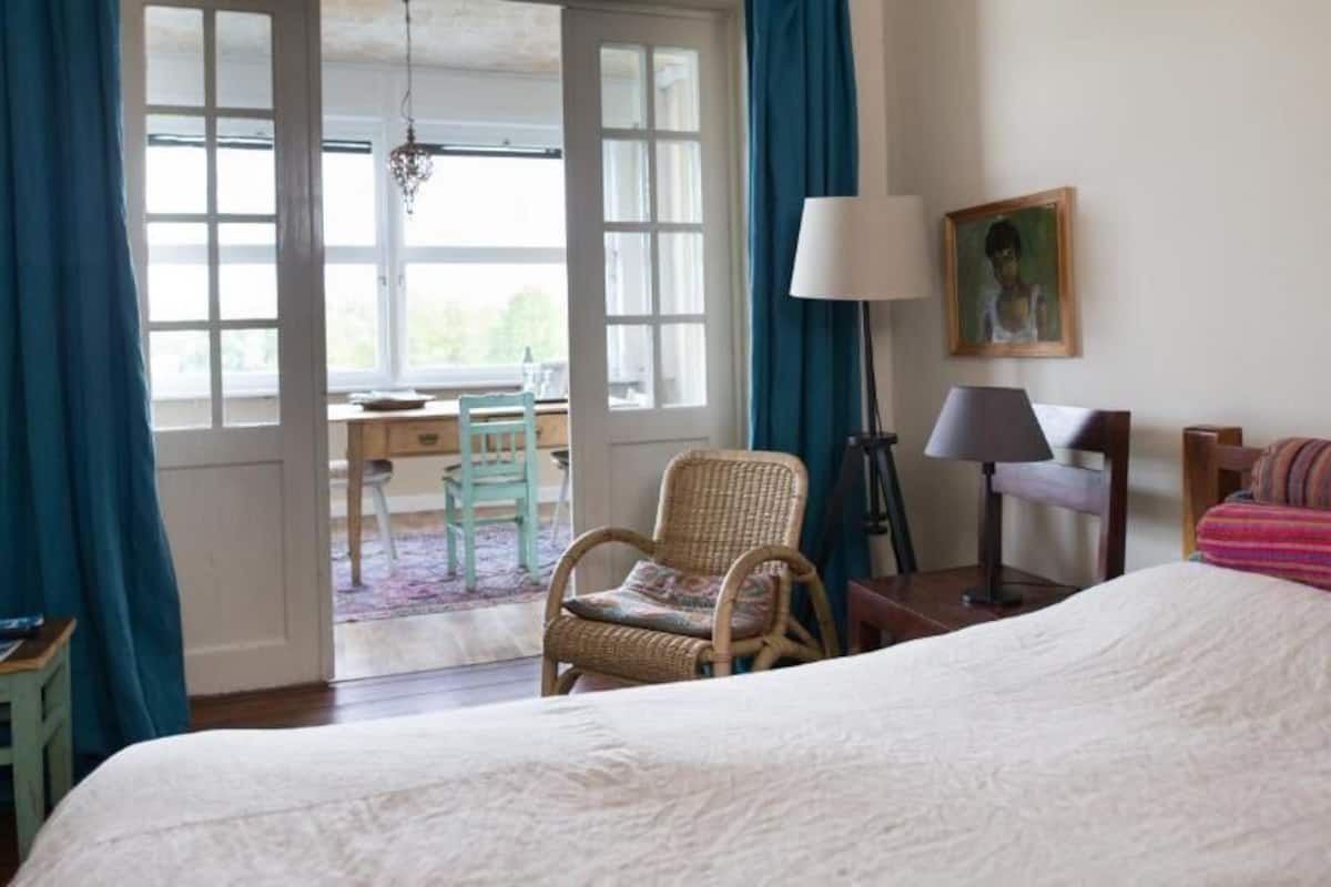 A bedroom with a large bed, a wicker chair, a desk with a chair, a lamp, and a painting on the wall.